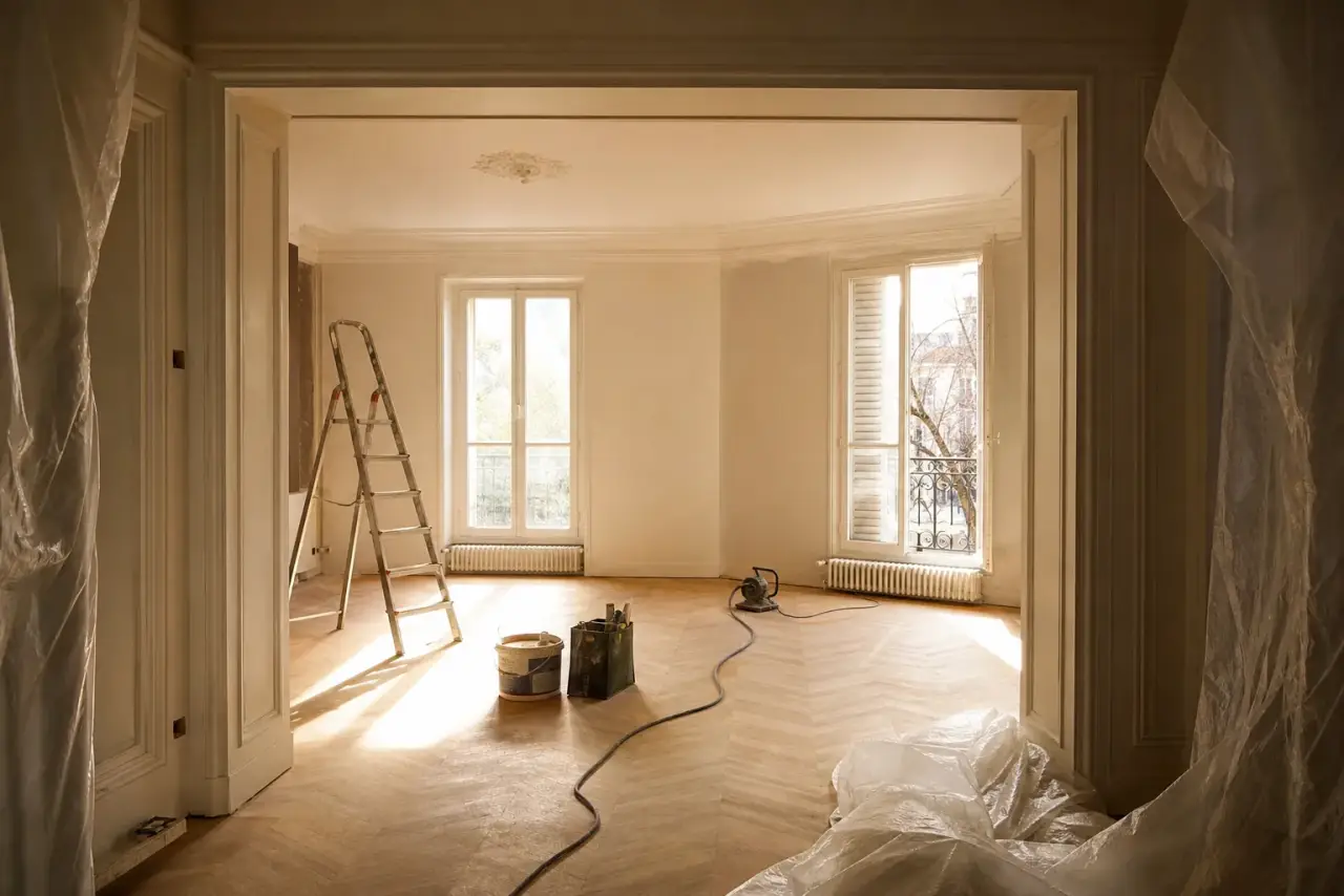Séjour d'appartement ancien en cours de rénovation, parquet poncé, moulures conservées, grandes fenêtres laissant entrer une lumière dorée de fin de journée, bâches et escabeau, photographie réaliste